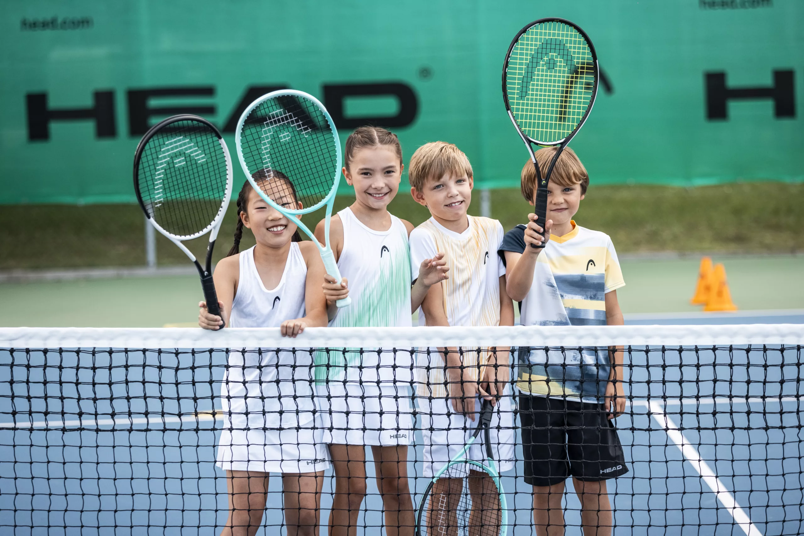 2 Girls and 2 Boys holding HEAD Rackets above a tennis net on court.